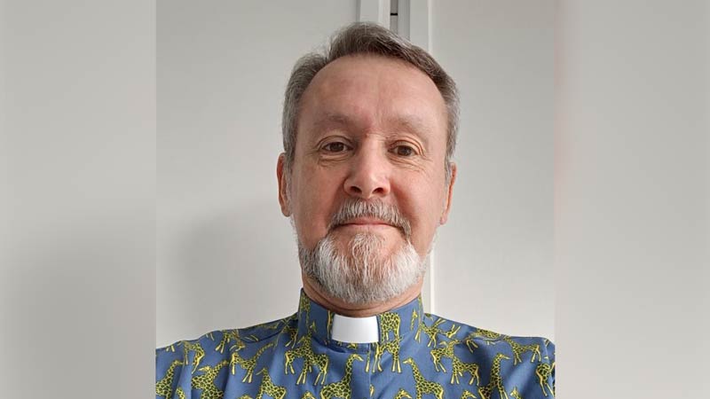 Patrick wears a clerical collar with a blue tunic decorated with a pattern comprising yellow giraffes