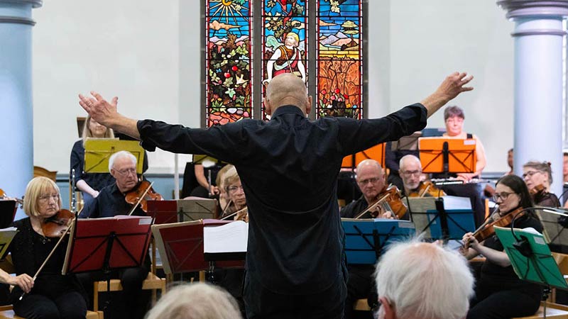 A conductor spreads his arms wide as he leads the orchestra in from of a colourful stained glass window depicting Jesus as the sower.