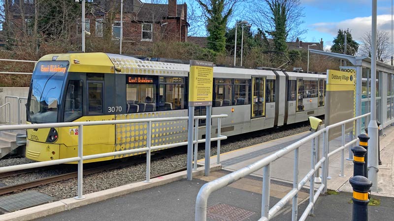 A yellow and gray Metrolonk tram pulls into the tram stop in Didsbury Village