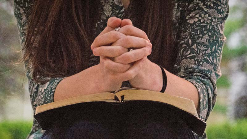 A person with long hair sitting outdoors, wearing a floral patterned shirt, clasping hands in prayer over an open Bible.