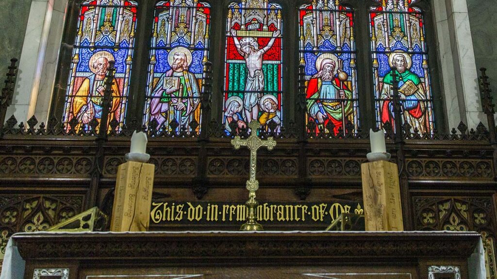 The altar at St James church beneath a stained glass window. Behind are the words 'This do in remembrance of me' gilded on oak paneling.