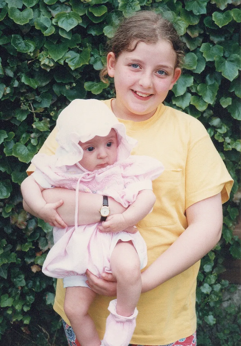 A young Rosalind is wearing a pale yellow t-shirt as she holds her baby sister