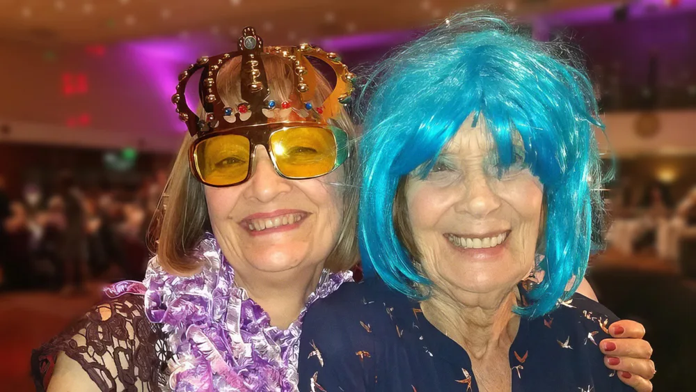 A smiling daughter and her elderly mother enjoying a party. The daughter is wearing a golden paper crown and her mother is wearing an electric blue wig.